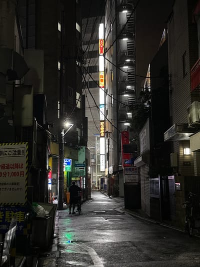 This image captures a narrow, dimly lit alleyway in Adachi, Japan, at night. The scene is wet, suggesting recent rain, with reflections visible on the asphalt. The alley is lined with multi-story buildings, typical of a Japanese urban environment, displaying a mix of businesses and residences.
Several illuminated signs are prominent, advertising various establishments. One sign, in Japanese, reads "椎橋歯科クリニック" (Shiihashi Dental Clinic), with its operating hours and contact information visible. Another sign points to a "LAWSON" convenience store, indicating it's 10 meters away. Other neon and illuminated signs for shops and services, though partially obscured and in Japanese, add to the bustling, commercial feel of the street.
In the foreground, a black and yellow sign with Japanese text warns "車にご注意!!!" (Beware of cars!!!), advising a fee of "1,000円 for every 10 minutes" after a certain time. There is also a sign that says "駐車" (Parking) and thanks the viewer.
A person is visible in the mid-ground, silhouetted against the lights, pushing a bicycle. The overall atmosphere is one of a typical urban night scene, with the interplay of light and shadow creating a slightly mysterious mood. The presence of the convenience store and dental clinic suggests a mix of residential and commercial activity. The time of day is clearly night, and the wet ground indicates recent rain.