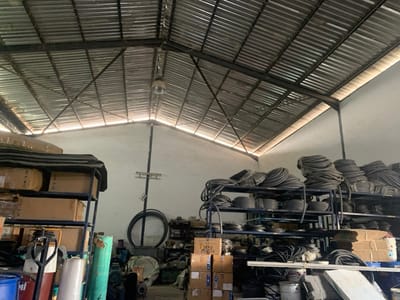 An interior view depicts a storage or workshop facility in Babawa, Nigeria. The scene is illuminated by natural light entering through gaps between the upper walls and the corrugated metal roof, which is supported by exposed steel trusses. A single industrial light fixture is suspended from the roof structure towards the center. The walls are painted white.
On the right side of the image, multiple dark-colored metal shelving units are densely populated with numerous coiled bundles of black flexible tubing or hoses, stacked in orderly rows. Towards the left, another metal shelving unit holds cardboard boxes, some displaying partial branding like "TOPO PAN." The floor area beneath and around the shelves is occupied by a variety of objects, including a red and black pallet jack, several buckets (one blue, others white), rolls of light-colored material, and various pieces of industrial equipment such as electric motors and pumps. Additional loose items and building materials are scattered on the floor. No individuals are present in the visible area.