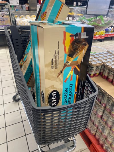 The image depicts a shopping cart filled with several large boxes of "Jamón Serrano" (cured ham). The boxes are decorated with Spanish flags and images of ham. In the background, shelves of canned drinks are visible, along with some supermarket displays. The flooring is tiled. The shopping cart is positioned in an aisle of a grocery store or supermarket. The overall scene suggests a retail environment in Spain.