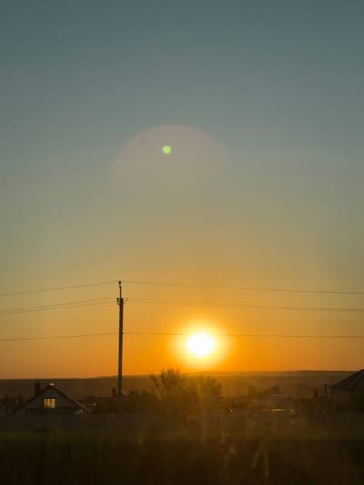 Captured from Bol'shiye Klyuchishchi, Russia, the landscape unfolds under a vibrant sunset. The sun, a luminous orange orb, dominates the sky, partially obscured by a cluster of trees and silhouetted buildings on the horizon. Overhead power lines cut across the sky, intersecting with a utility pole in the foreground. In the distance, a dwelling with a distinct gable roof is illuminated by a soft glow. The sky transitions from a light blue hue to a gradient of orange as it approaches the horizon. A lens flare is visible in the upper left quadrant of the image.