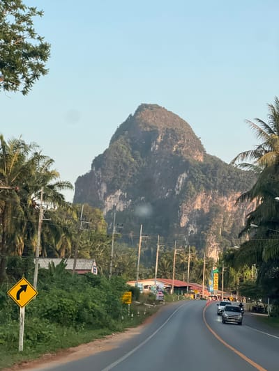 The image shows a road scene in Ban Nai Sa, Thailand, with a large, dominant mountain in the background. Palm trees are present along the roadside. An automobile is visible on the right side of the road. There are utility poles and wires on the side of the road. A yellow traffic sign indicating a left turn is visible in the foreground. There are buildings and signage along the roadside. The sky is light blue, suggesting daylight.