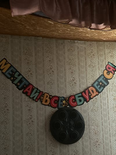 The image shows a banner with Cyrillic text that reads "МЕЧТАЙ! ВСЕ СБУДЕТСЯ!" which translates to "Dream! Everything will come true!". The banner is curved and made of colorful, individual letter cutouts, some of which are decorated with stars. Below the banner is a black, circular object with multiple smaller circular openings, resembling a speaker or a decorative element. The background is wallpaper with a subtle floral pattern. The top portion of the image shows a wooden surface and pink fabric, possibly part of a curtain or bedspread. The overall scene appears to be an indoor decoration. The image was taken in Saran, Kazakhstan.