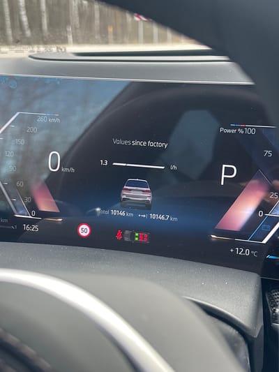The image captures a close-up view of a digital instrument cluster inside a modern car, likely from the driver's perspective. The car appears to be stationary or parked, indicated by the "P" (Park) gear selection and "0 km/h" on the speedometer.
The display shows various pieces of information:
- **Time:** 16:25
- **Outside Temperature:** +12.0 °C
- **Power:** "Power % 100" is displayed, possibly indicating maximum available power or battery charge.
- **Mileage:** The total odometer reading is 10146.7 km, with "Total 10146 km" also visible.
- **Fuel/Efficiency:** Under "Values since factory," a reading of "1.3 l/h" (liters per hour) is shown, likely indicating fuel consumption while stationary or idling, typical for some hybrid or electric vehicles.
- **Speedometer:** A digital speedometer shows "0 km/h" with a scale extending up to 260 km/h.
- **Warning/Indicator Lights:** A red circular sign with "50" suggests a detected speed limit. A top-down graphic of a car displays green indicators at the front and red at the rear, possibly for parking sensors or collision warnings. A pedestrian warning symbol is also visible.
Part of a steering wheel is visible on the right side of the frame, and reflections on the screen indicate daylight. There are also noticeable dust particles on the dashboard surface below the screen. The scene takes place in Mežezeri, Latvia.