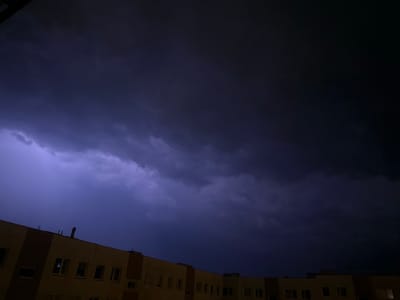 A building, likely residential or commercial, is visible in the lower portion of the image. The building's facade is predominantly light brown with darker brown accents. Several windows are discernible, some with reflections, and a chimney or ventilation pipe extends from the roof. Above the building, a dark, stormy sky dominates the scene. The clouds are dense and heavy, tinged with purple and blue hues, suggesting an approaching thunderstorm or active lightning. A faint, purple-blue flash of lightning is visible on the left side of the frame, illuminating the clouds from within. The overall atmosphere is dramatic and foreboding. The location is Świdnica, Poland.