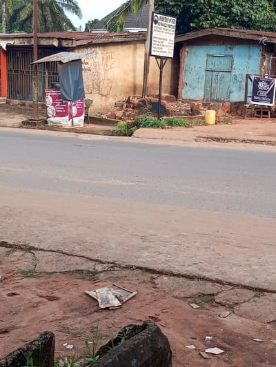 The image depicts a static roadside scene in Oko Polytechnic, Oko Town, Anambra State, Nigeria. The foreground consists of reddish-brown dirt and cracked concrete, with a crumpled, discarded object and small debris on the ground. A paved asphalt road, free of vehicles, occupies the mid-ground.
Along the right side of the road, a dirt shoulder fronts a series of single-story structures. From left to right:
1. A building with a reddish-brown, weathered exterior and a rusted corrugated metal roof, featuring a metal-grated entrance on its left side.
2. In front of this building, a temporary stall with a grey tarpaulin covering a corrugated metal awning stands on a white banner advertising "Primary Healthcare Services" and "Food Parties," displaying contact information. A green plastic chair is partially visible behind the stall.
3. A black metal signpost features a white sign for "MOUNTAIN OF FIRE AND MIRACLES MINISTRIES, SOUTHEAST REGION, R.D. 200 Oko Polytechnic, Anambra State," detailing service times and addresses.
4. Adjacent to the sign, a light blue, weathered building with peeling paint and a rusted corrugated metal roof shows exposed brickwork/debris at its base. It has a faded wooden double door.
5. A bright yellow plastic jerrycan rests on the dirt ground in front of the blue building.
6. On the far right, a black and white banner displaying "10" and additional text is visible, mounted on a white stand.
Lush green foliage, including palm trees, is visible behind the structures against an overcast sky. No individuals, actions, or interactions are present in the scene.