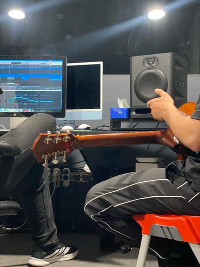 In a dimly lit room, possibly a recording studio, two individuals are present. One person, partially visible from the waist down, is seated in a chair and wearing black jeans and athletic shoes with white stripes. This individual is holding a guitar, with the neck and headstock clearly visible. The headstock is made of polished wood and features chrome tuning pegs. The other person, to the right, is also partially visible, with their hands near a speaker. They are wearing black pants with white piping.
The environment features electronic equipment, including a large computer monitor displaying a digital audio workstation interface with various tracks and waveforms. A second, smaller monitor is also present, as is a black studio monitor speaker. A blue box of tissues is placed on a shelf between the monitors and the speaker. The background is dark, with lighting fixtures visible on the ceiling. The floor appears to be carpeted. The scene suggests a music production or practice session. The image does not contain any specific visual cues that would allow for its location to be identified as Koghb, Armenia.