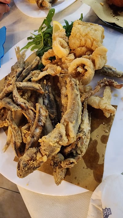 A close-up, top-down view captures a platter of fried seafood served in a restaurant setting in Latina, Italy. The platter, predominantly white, contains a generous portion of golden-brown fried calamari rings, small whole fish, and a larger piece of fried white fish, all coated in a crispy batter. A garnish of fresh green arugula and a lemon wedge is positioned on the left side of the platter. The food is placed on a brown paper liner. The background reveals a portion of another white plate with fried food and hints of the restaurant's white tablecloth.