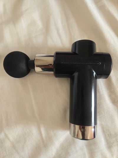 A black massage gun with a rounded black head and a silver tip rests on a white, wrinkled fabric surface. The massage gun is positioned horizontally, with the head on the left and the silver tip on the right. The fabric appears to be a bedsheet or similar textile. The lighting suggests an indoor setting. There are no people or any other objects visible in the image. The location in the city of Deira, United Arab Emirates is not discernible from the image itself.