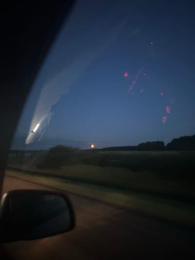 This media file captures a nighttime scene, likely from inside a vehicle, as suggested by the reflection on the windshield and the presence of a side mirror. The sky is a deep, dark blue, indicating it is either dusk or pre-dawn. There are several blurred, red lights in the sky, possibly distant aerial lights or reflections.
On the right side of the frame, a dark, silhouetted landscape with a defined edge runs vertically, suggesting trees or structures lining a road. A small, bright orange orb is visible in the distance against the dark landscape. The windshield reflects a bright streak of light, likely from oncoming headlights. The overall impression is one of travel through a rural area at night. The location context of Tyukalinsk, Russia, suggests a likely setting in a flat, open landscape typical of the region. No people are visible, and no text is present.