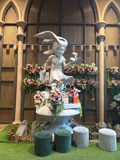 This vertical, eye-level medium shot captures a stone statue of a rabbit-eared figure with a flowing robe, holding a book and a small bird. The statue is adorned with colorful flower arrangements featuring roses, carnations, and foliage in shades of pink, red, and white. Several small rainbow flags are strategically placed around the statue, attached to a vertical white strip and also near the statue's base.
Behind the statue, a slatted wooden wall serves as a backdrop, with large decorative floral wreaths hanging from it. To the right of the statue, three rounded, quilted ottomans are arranged on a patch of artificial turf. Two are light gray, and one is a deep green. Beyond the ottomans, a naturalistic display of rocks and logs adds a rustic element to the setting.
The lighting appears to be artificial, casting distinct shadows that suggest an indoor environment. The overall atmosphere is festive and celebratory, likely related to a Pride event given the prominent display of rainbow flags. The visible text on the books held by the statue reads "Twilight Angel," "Giant's Secret," and "Witty Prince" in English, with some Thai text also present below. The image seems to be taken in a public or event space in Ban Na Chom Thian, Thailand.