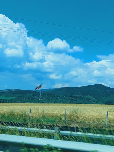 This media file depicts an outdoor scene that appears to be captured from a moving vehicle, given the slight blur and reflective elements on the right side of the image. The orientation of the image is vertical.
The primary subjects include a vast expanse of blue sky filled with large, fluffy white clouds. To the left, several dark power lines stretch diagonally across the frame. In the middle ground, a rolling landscape is visible. A hillside covered with dense green vegetation dominates the background. In front of the hillside, a field of golden crops, likely wheat or barley, stretches across the scene. A flagpole with a red and green flag is planted in this field, partially obscured by the crops. On the far right, a blurred section suggests a road or a reflective surface, possibly the window of the vehicle, with green foliage and vertical elements that might be guardrails or similar structures.
The scene appears to be set in the daytime, under clear to partly cloudy weather conditions, as indicated by the bright sky and prominent clouds. The presence of the golden fields and lush green hills suggests a rural or agricultural setting, consistent with the provided location context of Eleshnitsa, Bulgaria.
There is no discernible text within the image. The overall mood is serene and natural, capturing a moment of travel through a scenic countryside.