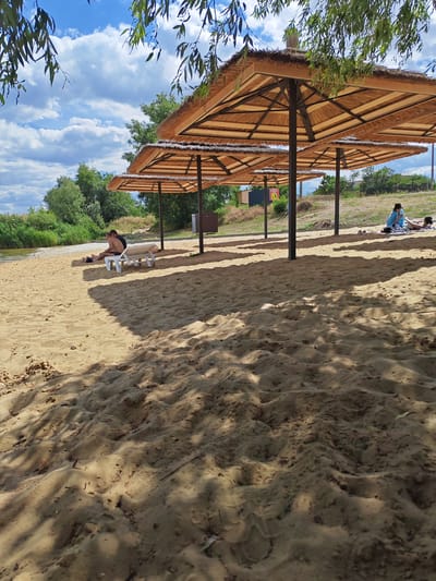 On a sandy beach next to a body of water, several thatched-roof umbrellas provide shade. In the foreground, the sand is textured with footprints and shadows. In the middle ground, two people are sitting on a white lounge chair under one of the umbrellas. Further back, another person is lying on the sand. The background shows a line of trees, a grassy slope, and a bright blue sky with fluffy clouds. The scene appears to be a recreational area, possibly a beach resort or park, located in or near the city of Belaya Kalitva, Russia.