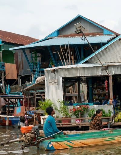 The image depicts a floating village in Siem Reap, Cambodia, specifically within the locality of Soutr Nikom. In the foreground, a narrow boat with an outboard motor is on the water. A man wearing a blue long-sleeved shirt and green shorts sits in the boat, holding an oar and looking upwards. To his right, a woman with dark hair and a patterned long-sleeved shirt sits facing forward, looking towards the right side of the frame. Behind the boat, a two-story stilt building with a blue corrugated metal roof is visible. The lower level of the building features a shop with shelves stocked with various items. A sign on the building reads "MAIS DRINKS & SNACKS EST 1988". A solar-powered light pole is positioned in front of the building. Other stilt structures with corrugated metal roofs and wooden supports are visible in the background, suggesting a dense, water-based community. The water is calm and reflects the overcast sky.