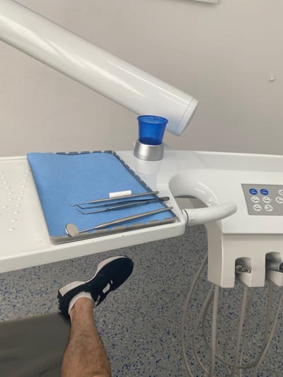 The image depicts a dental examination room. A white dental chair is partially visible in the foreground, with a person's leg wearing a black and white sneaker resting on it. A dental tray is placed on a white surface, holding a blue disposable mat and several dental instruments, including a mirror, tweezers, and a dental probe. A blue plastic cup sits on a silver base next to the tray. The background features a white wall and a white arm extending from an overhead light fixture. The floor is speckled blue and gray. This scene is located in a dental practice in Bad Nauheim, Germany.