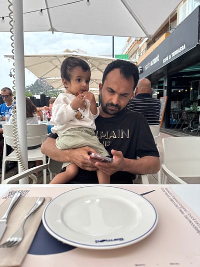The image depicts an outdoor dining scene on a restaurant terrace in L'estartit, Spain. In the foreground, an adult male with dark hair and a beard, wearing a black "BALMAIN PARIS" t-shirt, holds a young child. The male is looking down at a smartphone, held in his left hand, while the child sits facing forward, wearing a white long-sleeved top with a cartoon graphic and light green shorts, with a small, reddish-brown object (likely food) in its mouth and right hand.

On the table in front of the subjects, a light pink placemat features a QR code and partial text for "Barmoodeperda." An empty white plate with a blue rim and "Bermood" printed on it is centrally placed. To the left, a knife and a fork rest on a brown napkin.

The background shows other patrons and elements of the restaurant. White umbrellas provide shade over multiple tables and chairs. To the far left, a man with glasses in a blue shirt is visible. To the right, a bald man in a striped shirt is seated further back. A dark building exterior, marked "Restaurant" and "Cafe & Terrassa," forms part of the immediate background. Distant hills or mountains are visible under an overcast sky. Overhead, string lights are strung between the umbrella structures.