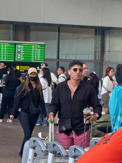 A group of people are standing in what appears to be an airport, waiting. One person is front and center wearing sunglasses, a black jacket, and patterned red and pink pants, holding a Starbucks drink and a luggage cart. Another person in a black outfit and beige baseball cap is slightly behind. An airport flight information display board can be seen on the left.