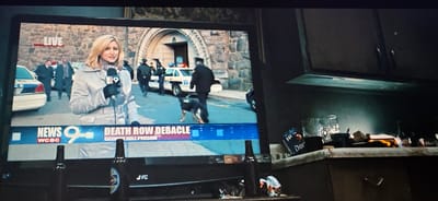 A television screen displays a news report, with a female reporter in the foreground holding a microphone. Behind her, a police presence is visible, including officers, a police car, and a police dog. The news banner at the bottom of the screen reads "NEWS 9 WCBC" and "DEATH ROW DEBACLE - GEORGE HILL PRISON". The television is situated in a dimly lit room, with kitchen cabinets visible in the background and bottles on a counter. The setting appears to be a domestic environment, rather than a public location in Esbjerg, Denmark.