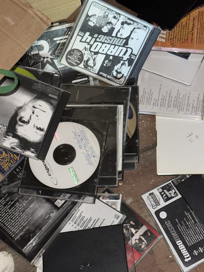 This media file is a top-down, close-up shot of a collection of CDs and their cases, strewn on a rough, dirty surface that appears to be the floor. The setting is dimly lit, suggesting an indoor location possibly in a basement or storage area, and the surface is cluttered with debris, giving it a neglected feel. The CDs themselves are varied, with some featuring colorful album art and others having handwritten labels.

Prominently displayed is a CD case with the bold text "TURBOMUSIC!" on a black background, featuring black and white images of people. Another CD case, also with "TURBO MUSIC!" branding, shows cartoonish figures. There are also several other CD cases, some with red and white designs, and a few with what appear to be Russian text. One CD has a handwritten label that includes "Metallika" and "Top (Mp3)". Another visible CD has the brand "esper" and text that reads "creating space to".

Among the visible text are track listings and artist names, including "LORINDO", "KIM CARNES", "WHIGFIELD", and "BAD BOYS BLUE". Some of the Russian text visible on album covers translates to song titles or artist names such as "ДУШИ", "ЗВЕЗДНАЯ СЕСТВИЯ", and "РУССКИЙ". The overall impression is a disorganized pile of music media, possibly in storage or discarded. The presence of Russian text indicates the origin or distribution within a Russian-speaking region. There are no discernible people actively engaged in an activity, but rather a static arrangement of media items.