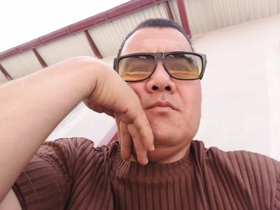 A close-up, selfie-style shot captures a man with dark hair and fair skin, wearing dark-rimmed sunglasses with yellow lenses and a brown ribbed t-shirt. He rests his right hand on his chin, with his left arm and shoulder visible. The background features a portion of a light-colored building and an outdoor roof structure made of corrugated metal with a burgundy trim. The lighting suggests an outdoor, daytime setting. The location is identified as Juma, Uzbekistan.