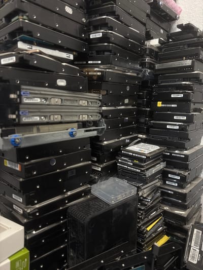 The image depicts a dense, indoor storage area filled with stacks of computer hard drives. The hard drives are primarily black and appear to be of various sizes and models, with some featuring labels and barcodes. They are arranged in precarious towers, with some leaning and others securely stacked. In the foreground, there are metal trays with blue fasteners, suggesting these might be part of a server rack or storage system. To the left, partially visible, are boxes, one white and one green, with some text on them. To the right, a black, ventilated electronic device is present, possibly a power supply or server component. A clear plastic container with what appears to be small electronic components is also visible. The overall impression is one of a disorganized or densely packed electronic component storage facility. The location is identified as Mülheim an der Ruhr, Germany. No people are visible in the image.