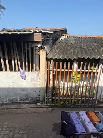 In Moratuwa, Sri Lanka, a scene unfolds beneath a clear blue sky. The foreground displays a clothesline with four articles of clothing, two dark, one purple, and one pink with blue flowers, secured by green clothespins. Behind the clothesline, a weathered fence made of vertical wooden slats, some stained brown and others white, partially encloses an area. A green vine with broad leaves climbs the fence. Beyond the fence, the roofs of two structures are visible. The structure on the left has a corrugated roof and a white brick wall below, with vertical wooden slats forming a barrier. A purple and white patterned cloth hangs from these slats. The structure on the right also features a dark, corrugated roof and a fence of similar wooden slats. The overall environment suggests a residential area with simple construction.