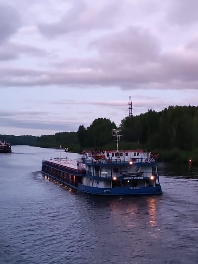 The image captures a wide river or canal in Aleksandrovskoe, Russia, under an overcast sky with purple-tinged clouds. A large blue and white barge, named "ИНЖЕНЕР ШУХОВА" and "ТАГАНРОГ", is the main subject, traveling along the waterway. Its lights reflect on the water's surface, creating streaks of light.
In the background, a line of dense green trees borders the waterway. A tall metal transmission tower is visible behind the trees, to the right of the barge. Another vessel is further in the background. A channel marker is visible along the right shoreline. The water appears slightly choppy.