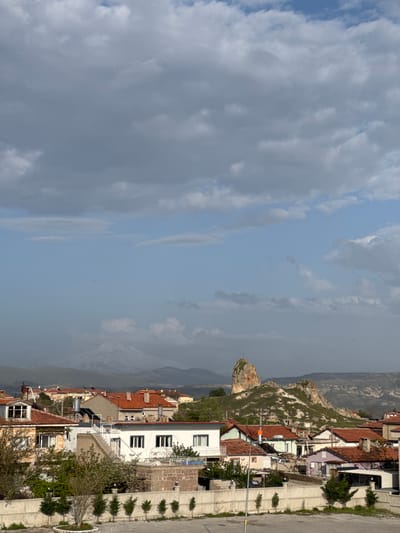 The image presents a view of a hillside town under a partly cloudy sky. In the foreground are the roofs of closely spaced buildings, predominantly red-tiled. A notable rocky outcrop rises behind the buildings, connecting the town to the mountainous landscape in the distance. The architecture features a mix of traditional and modern elements. Adjacent to the buildings is a paved road with regularly spaced, small trees. The town is located in Ürgüp, Turkey, known for its unique geological formations and historical sites.