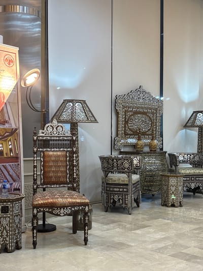 Elaborately inlaid furniture pieces are displayed against a white wall. The items, decorated with intricate patterns and light-reflecting materials, include chairs with lamps, tables, and a framed piece with Arabic calligraphy. The furniture is arranged in a row, showcasing detailed craftsmanship and reflective surfaces. The background is a modern interior with sleek surfaces.