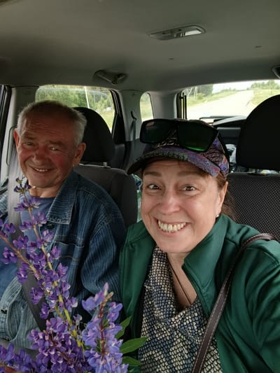 Two individuals are seated in the front seats of a car, facing the camera. The person on the left, an elderly man with grey hair, smiles broadly and is wearing a blue denim jacket over a blue t-shirt. He is holding a bouquet of purple lupine flowers. The person on the right, a woman with brown hair, is wearing a green jacket over a patterned scarf and a baseball cap with sunglasses perched on top. She also smiles at the camera. The interior of the car is visible, including the headrests and the ceiling. Outside the car, through the windows, a rural road lined with trees can be seen, suggesting a location in the countryside, possibly near the village of Gribchikha, Russia, given the context. The overall impression is a candid, happy moment shared between two people on a journey.