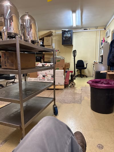 The image depicts a cluttered storage room or workshop area, likely within the Bnei Shimon Regional Council, Israel. In the foreground, a person's legs and feet, clad in grey pants and brown shoes, are visible, suggesting they are sitting or standing, possibly near a metal shelving unit.

The shelving unit, made of stainless steel, occupies a significant portion of the left side of the frame. It is laden with various items, including large, silver, dome-shaped serving vessels on the top shelf, and boxes of what appear to be food products or supplies on lower shelves. Some of these boxes have text that is partially visible, with some of it appearing to be in Hebrew.

The environment is characterized by a yellow-painted wall and a light-colored ceiling with fluorescent lighting. Various objects are scattered throughout the room, indicative of a working or storage space. These include stacks of cardboard boxes, some labeled with brand names like "Knorr," and a black server rack with visible wiring mounted on the wall. A red and black manual pallet jack stands near a stack of pallets and boxes. A black office chair is positioned in the mid-ground, and to its right, a white shelving unit holds various items, including what appears to be jackets. A large black trash bin with a purple trash bag is situated in the right side of the image. The floor is a mosaic of speckled light-colored tiles and areas with a different, more textured pattern. The overall impression is one of a functional but disorganized space.
