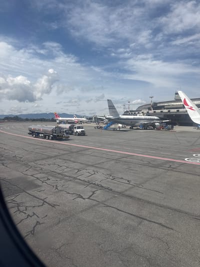 The image depicts an outdoor scene at an airport in Rionegro, Colombia, likely José María Córdova International Airport, given the context of Avianca aircraft. The foreground shows a cracked asphalt surface with painted red and white lines, suggesting it is part of a taxiway or tarmac. In the mid-ground, two large silver fuel trucks are parked. The truck on the left is a tanker truck with multiple axles, and a smaller white truck is positioned behind it. Beyond the trucks, several Avianca airplanes are visible. One large airplane, labeled "AVIANCA-COLOMBIA," is parked with stairs leading to its entrance, and several individuals are standing near it. Another Avianca airplane, with a red and white tail, is parked further in the background. In the far background, a modern airport terminal building with large windows and a curved roof is situated. Mountains are visible in the distance, shrouded in clouds. The sky is partly cloudy with blue patches. The overall scene captures a moment at an active airport, with ground support vehicles and aircraft present.