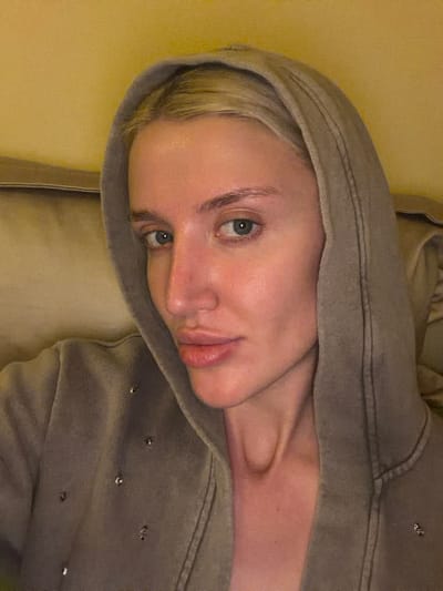 The image features a close-up portrait of a person, likely female, with fair skin and blonde hair, looking directly at the camera with a neutral expression. The person's head is tilted, and their light-colored eyes appear greenish-blue. Their lips are slightly parted.
They are wearing a hooded sweatshirt of a muted khaki-grey or taupe color, with the hood pulled up to frame their face. Small, silvery metallic embellishments, possibly studs or sequins shaped like tiny flowers or stars, are visible on the fabric of the sweatshirt, particularly on the right side of the image. The fabric of the garment shows a soft, textured quality. The person's neck and collarbone are visible, showing natural contours.
The background is indistinct, consisting of a plain yellowish or light beige surface, suggesting an indoor setting, possibly a wall or upholstered furniture. The lighting appears dim and soft. There is no visible text in the image. The scene is set in Nerin, Montenegro.
