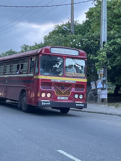 A red bus with a yellow stripe and white lettering is driving down a paved road. The bus has "LANKA ASHOK LEYLAND" written on the front in white letters. The license plate reads "NB 8405". There are trees and utility poles on the right side of the road. The sky is overcast. The bus is likely in Koggala, Sri Lanka, given the script on the bus. There are no people clearly visible in the foreground or on the bus's interior, though figures can be seen through the windows.