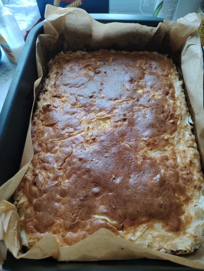 A close-up overhead view shows a rectangular baked good, likely a cake or pie, resting in a dark grey baking pan lined with brown parchment paper. The baked item has a golden-brown, slightly uneven surface with some areas appearing lighter and others darker, suggesting variations in baking. The edges of the baked good are visible where they meet the parchment paper, and appear to be made of thin, layered pastry or dough. The background is blurred and shows some white and dark objects, but provides no clear context. There is no indication of the location in Vladimirovka, Russia.