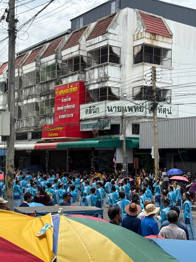 In Prakhon Chai District, Thailand, a crowd of people, many dressed in blue traditional clothing, are gathered on a street. The scene is dominated by a multi-story building with a weathered facade, featuring red-tiled roofs and balconies with metal grates. Red and green awnings extend from the lower floors, covering shopfronts. Several signs in Thai script are visible on the building. Overhead, a dense network of electrical wires crisscrosses the sky. In the foreground, parts of brightly colored umbrellas are visible, suggesting a public event or market. The people in the blue attire appear to be part of a procession or performance, with some holding small flags or banners. The atmosphere suggests a cultural celebration or festival.