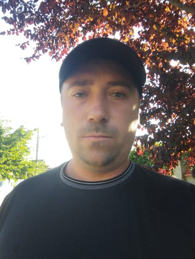 A close-up selfie of a man wearing a black baseball cap and a black t-shirt with a grey patterned neckline. He has light skin, short brown hair, and stubble. The background is out of focus, showing bright sunlight filtering through the red-brown leaves of a tree. To the left, blurred green foliage and a utility pole are visible. The overall lighting suggests it is daytime, possibly late afternoon given the angle of the sun. The image appears to have been taken outdoors.