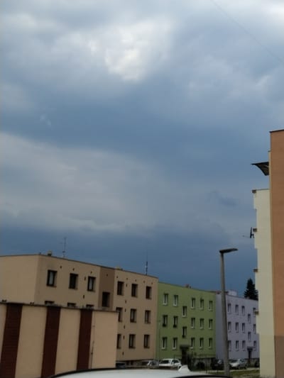 The image depicts a residential street scene in Hluboká nad Vltavou, Czechia, under a cloudy sky. In the foreground, the top of a white car is visible. Adjacent to it, a building with a light brown facade and vertical dark brown stripes is partially visible. Further back, a beige apartment building with multiple windows is lined up. To its right, a light green apartment building with numerous windows is followed by a light purple apartment building, also with many windows. A tall, grey lamppost stands in front of the purple building. In the background, a dark green coniferous tree is visible behind the apartment buildings. The sky is filled with dark, heavy clouds.