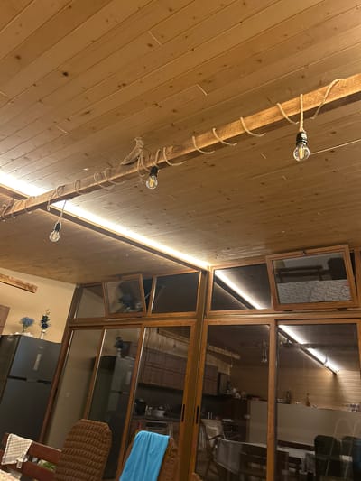 This image depicts the interior of a room with a wooden ceiling. Three exposed filament light bulbs are suspended from a wooden beam by rope, with additional rope wrapped around the beam. A strip of LED lighting runs along the ceiling. A large glass window structure with wooden frames dominates the lower portion of the image, revealing a glimpse of a kitchen or dining area with wooden cabinetry and a refrigerator. On a table in the foreground, there are two chairs, one with a brown textured seat and the other with a blue cloth draped over it, and a checkered cloth. The overall aesthetic suggests a rustic or bohemian interior design. There are no people visible in the image. The location is identified as Kamenar, Bulgaria.