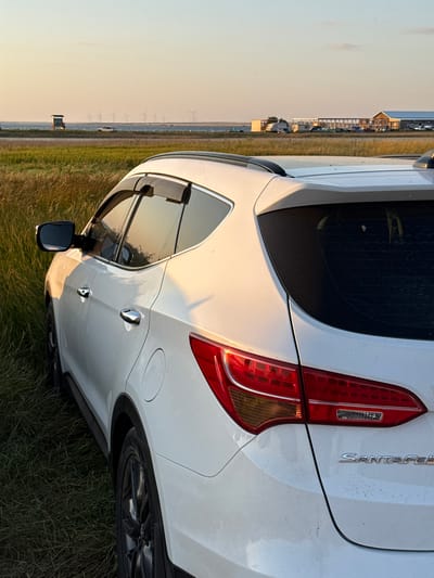 The image shows the rear portion of a white SUV, identified by the "SANTA FE" badging, parked on a grassy field. The car is facing right in the frame, with its rear left tire and bumper visible. The sun is low in the sky, casting a warm, golden light that illuminates the car and the surrounding landscape. The sky is a soft gradient of orange and yellow, indicating either sunrise or sunset. In the background, a row of wind turbines stretches across the horizon, suggesting a rural or open area. A few structures, possibly buildings or sheds, can be seen in the far distance, along with what appears to be a road and some vehicles. The overall scene evokes a sense of tranquility and openness. The image is oriented vertically, with the car's rear facing the top of the frame. There is no text visible on the car other than the model name. The weather appears clear and calm.