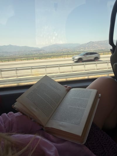 A close-up shot shows a person reading a book while traveling. The book is open and held by the person's hand. The pages of the book display text in Cyrillic script. In the background, through a window, a highway with a car in motion is visible, leading towards a landscape of hills and mountains under a clear sky. The scene appears to be captured inside a vehicle, possibly a bus or car, as indicated by the window and the presence of a car headrest. The light suggests it is daytime. The location is described as being in the city of Efeler, Turkey.