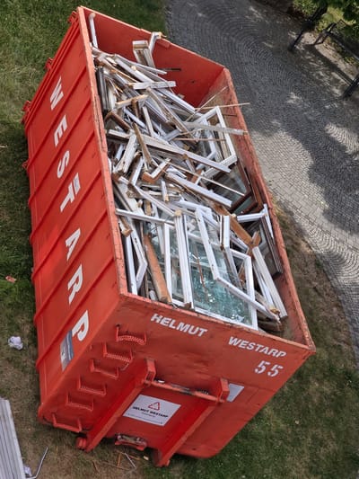 The image shows an aerial view of a large, orange dumpster filled with dismantled window frames and broken glass. The dumpster is situated on a grassy area next to a paved walkway made of cobblestones. The dumpster has white lettering on its side that reads "WESTARP" vertically and "HELMUT WESTARP Entsorgung 55" horizontally. There is a recycling symbol with a triangle and arrows on a white label on the side of the dumpster. The scene appears to be outdoors, possibly in a residential or construction area, given the presence of the dumpster and the surrounding grass and paving. The time of day seems to be daytime, as indicated by the shadows cast by the trees and the overall lighting of the scene. The location is Aschaffenburg, Germany.