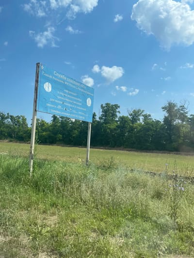 This media file depicts an outdoor scene on a bright, sunny day. The sky is a vivid blue, dotted with fluffy white clouds, suggesting fair weather. The foreground is dominated by a tall, blue sign with white text and graphics, mounted on two metal posts. The sign appears to be weathered, with some peeling paint.
The text on the sign, written in Russian, indicates it is from the "Emergency Service '01'" and the "Main Directorate of the Ministry of Emergency Situations of Russia for the Republic of Kalmykia." It informs the public about what to do in case of emergencies, providing emergency numbers like "01," "112," and a trust line number. Two circular graphics, resembling stylized basketballs or target symbols, are also visible on the sign.
Behind the sign, a strip of lush green grass and vegetation stretches across the frame, with some taller plants and wildflowers interspersed. Further in the background, a dense line of trees forms a natural backdrop, suggesting a rural or park-like setting. The trees are a vibrant green, indicating they are in full foliage. The overall impression is one of a functional public information sign in a natural environment, likely providing important safety information to those in the area. The orientation of the image is portrait, with the sign appearing prominently on the left side.