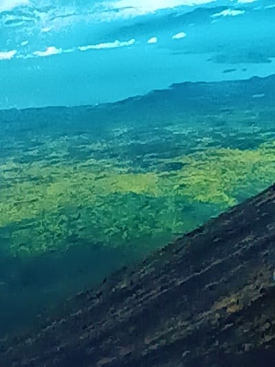 An aerial view captures a landscape dominated by green and brown terrain. The sky, a light blue with scattered white clouds, contrasts with the darker hues below. A distant mountain range lies along the horizon, partially obscured by a blue haze. The textured foreground suggests a rugged, uneven slope.