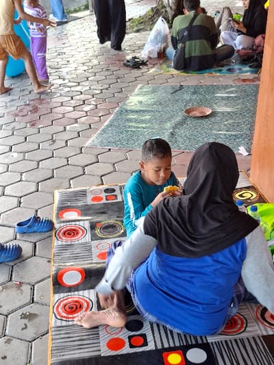 The scene is set on a paved area in Kecamatan Tulung, Indonesia, with a woman in a black headscarf seated on a black and white checkered mat, wearing a blue shirt. She is facing towards the left and slightly bent over. Her bare right foot is visible. In front of her sits a young boy wearing a blue shirt and blue pants. He is looking down and eating something, possibly corn. The boy is positioned on the left side of the mat, facing the camera. There is a blue plastic container with a blue cap, and a green and yellow container to the right of the woman. Behind the seated woman is a blue mat with a lighter blue patterned design, lying diagonally to the right. There are several people visible in the background: a young girl in a purple dress standing on the left side of the image; a woman wearing a black hijab is sitting with a man dressed in a dark green long-sleeved shirt, both on the right side of the image; and a person wearing black clothing is partially obscured by a building on the far right of the image. The paved area is made of hexagonal tiles. A tree with thick branches extends from the top right side of the image, reaching towards the left side. The background suggests a public area with possible retail businesses or services nearby. The scene appears to be a public area outside, perhaps a market or park, with people gathered for a gathering or social event.