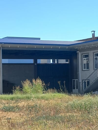 The image shows an outdoor scene with a building and overgrown vegetation. The building appears to be a commercial or industrial structure with a blue roof and gray walls. There are windows and a staircase visible on the right side of the building. In the foreground, there is a field of dry and green grass with some taller plants. The sky is clear and blue. The scene is likely located in the city of Yorkatay, Uzbekistan.