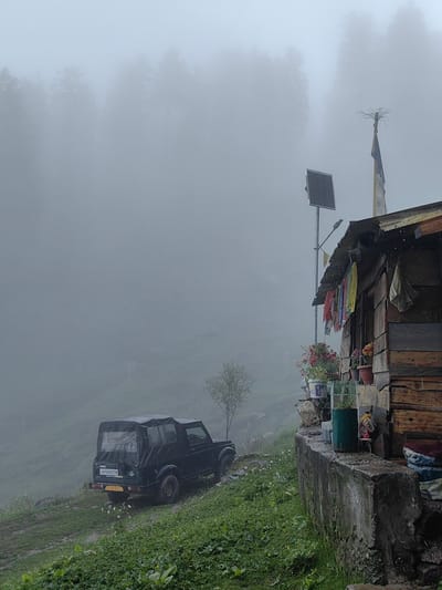 In Aleo, India, a dark green convertible jeep is parked on a muddy, grassy hillside. The jeep has a soft top and a license plate visible. To the right of the jeep is a rustic wooden structure with a corrugated metal roof, adorned with prayer flags and other colorful fabrics. A solar panel is mounted on a pole next to the structure, and potted plants, including red and pink flowers, are placed on a stone ledge. The background is obscured by heavy fog, revealing the silhouettes of trees in the distance. The ground appears wet, suggesting recent rain.