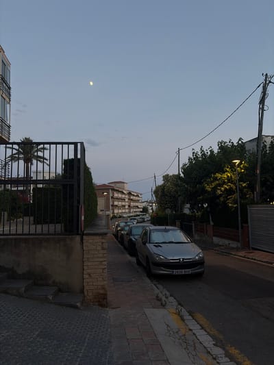 This image captures a street scene in Cambrils Badia, Spain, likely during the late afternoon or early evening due to the soft, ambient light and the presence of the moon in the sky. The scene is oriented vertically, with a street running along the right side of the frame.

Several cars are parked along the street, including a silver Peugeot with the license plate "2836 BHX". A street lamp is illuminated, casting a warm glow on the surrounding trees, which appear full and leafy. Buildings line the street, some with balconies. A black metal gate is prominent in the foreground, with a palm tree visible behind it. The ground is paved with a combination of bricks and stones, with a yellow line painted along the edge of the road.

The sky is a pale blue, with a faint, full moon visible in the upper left quadrant of the image. The overall atmosphere appears calm and tranquil, typical of a residential area at dusk. There are no people visible in the frame.