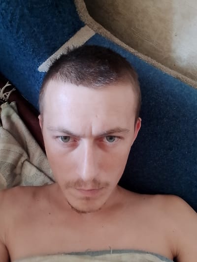 The image is a close-up, top-down selfie of a young man with short brown hair, light skin, and green eyes. He has a faint mustache and a somewhat furrowed brow. He appears to be lying down, with his upper body visible, covered by a blanket or sheet. The background consists of a textured blue surface, possibly a rug or upholstery, and a wooden surface. The image was taken in a home environment.