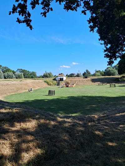 In the city of Brill, Netherlands, a semi-circular grassy area is visible, bordered by a dry, reedy embankment. A small, white building with a thatched roof sits on the embankment, featuring a wooden staircase leading to its entrance. A playground structure, resembling a ship, is located in the center of the grassy area. A dark wooden bench is positioned on the embankment to the left, while two picnic tables and a dark wooden cube are scattered across the grass. The scene is framed by the dark silhouettes of tree branches against a bright blue sky.