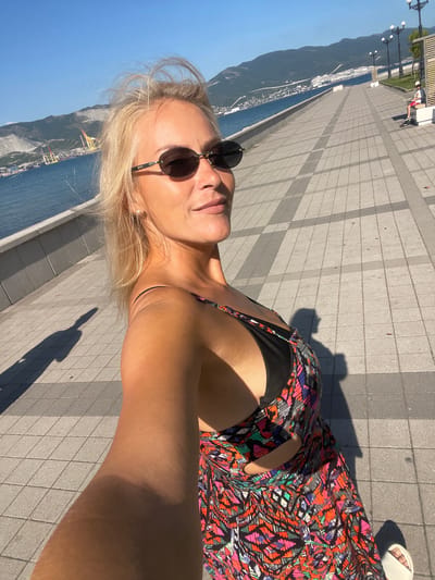 This is a selfie taken by a blonde woman with sunglasses and a colorful sundress. She is on a paved walkway next to a body of water, with mountains in the background. The sun is bright, casting shadows on the walkway, suggesting it is daytime. The woman appears relaxed and happy, looking towards the camera with a slight smile. In the distance, there are buildings and what appears to be a port or industrial area. The overall scene evokes a sense of leisure and a vacation atmosphere.